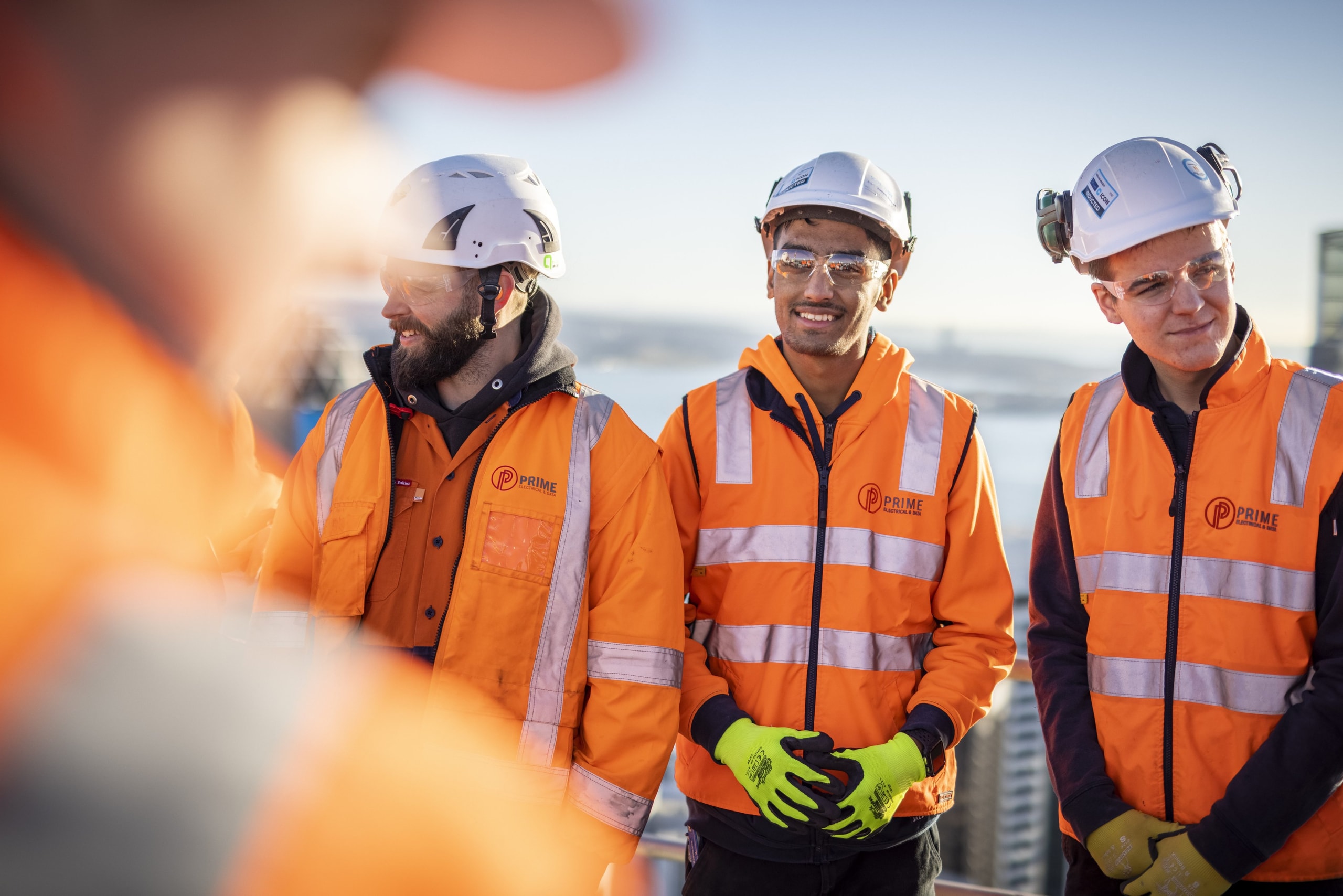 Three Prime Electrical & Data electricians in hi-vis and hard hats on a high-rise rooftop.